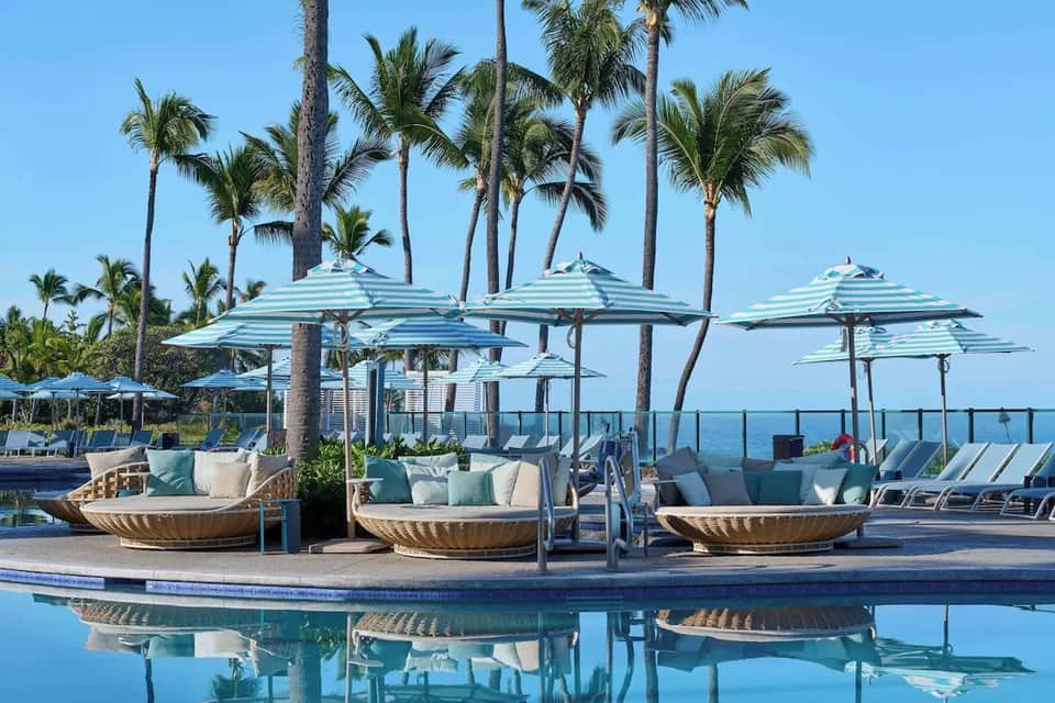 Luxury poolside lounge area with turquoise striped umbrellas, shell-shaped seating pods, and palm trees with ocean view