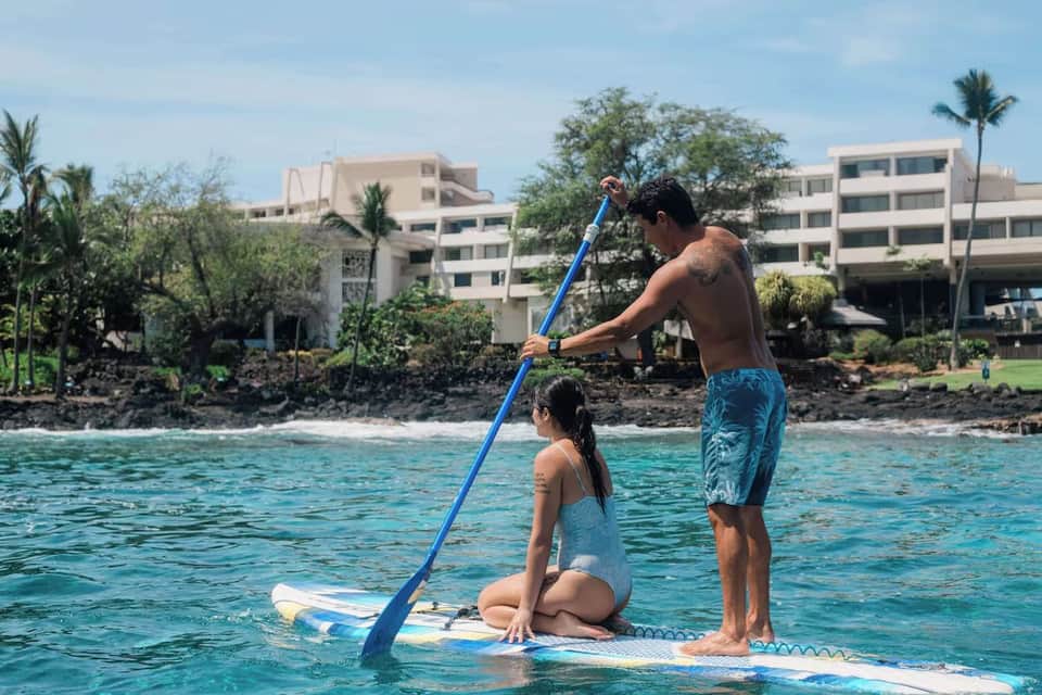 Two people paddleboarding on turquoise water near coastal resort buildings and palm trees