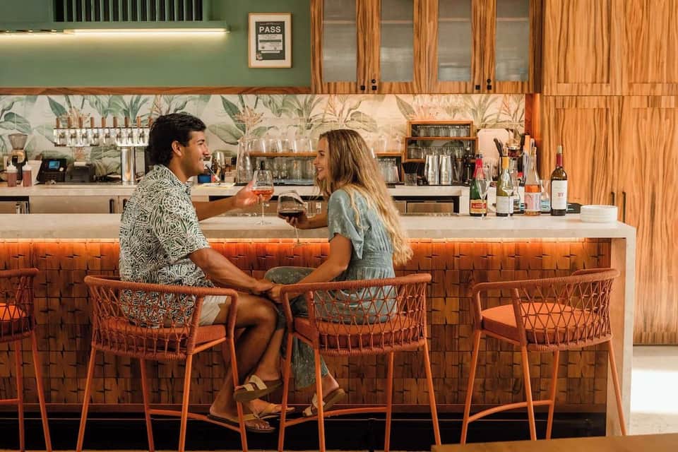 Stylish resort bar interior with couple at counter, orange woven chairs, and tropical botanical backdrop