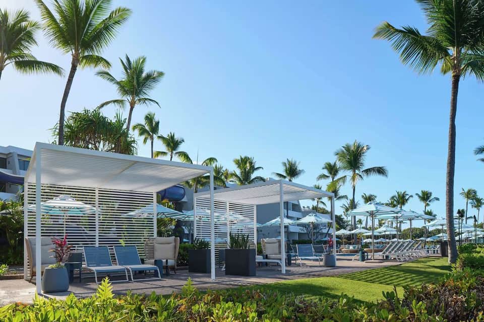 Modern beachfront resort pathway with white lounge chairs, palm trees, and ocean view under clear sky