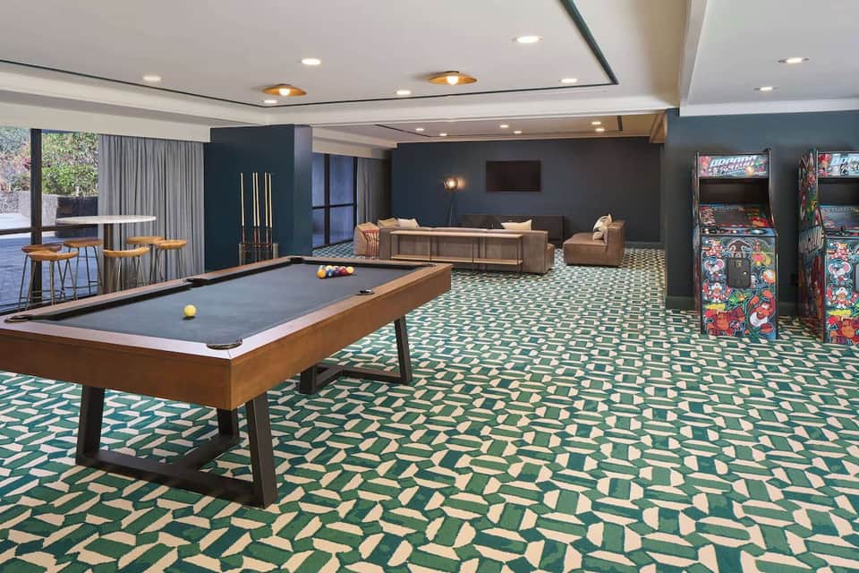 Resort game room with billiards table, arcade games, lounge seating, and geometric patterned carpet