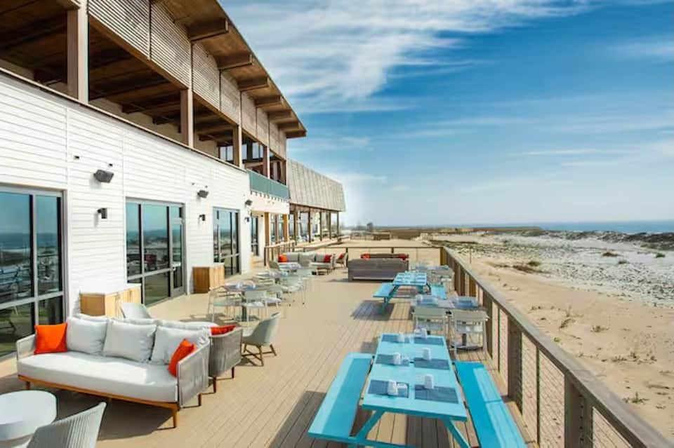 Coastal deck with lounge seating, dining tables, white building, and expansive beach landscape