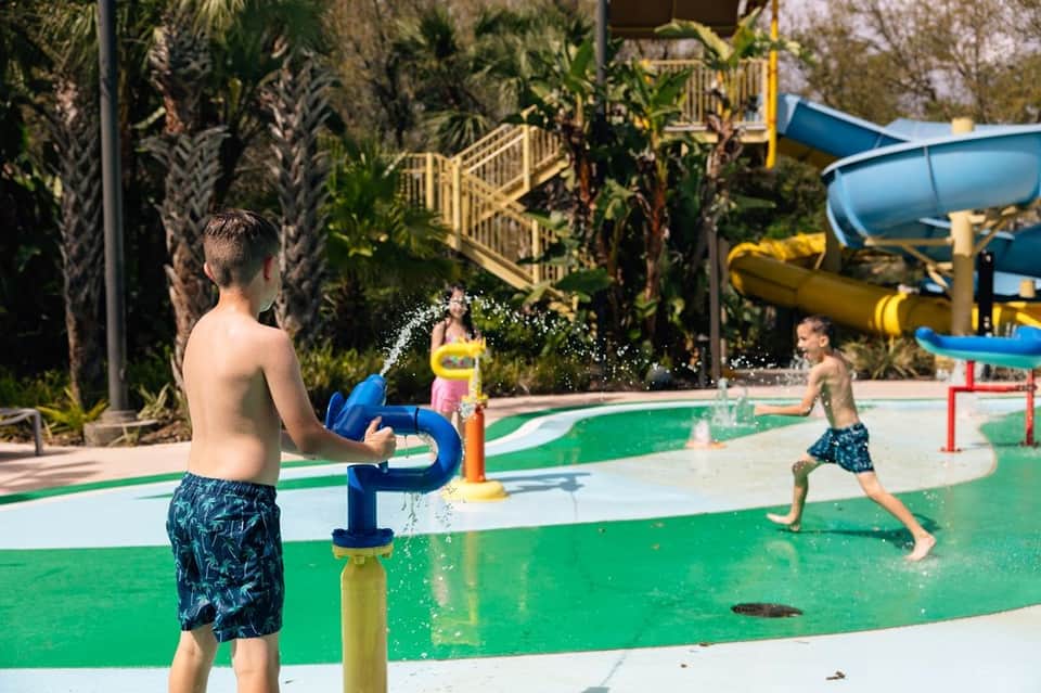 Family water park with splash pad features, water jets, blue and yellow water slides, and palm tree landscaping