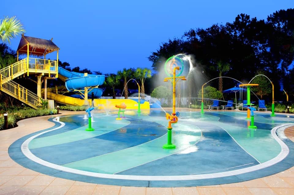 Illuminated evening aquatic play area with colorful water features, interactive jets, and palm tree surroundings