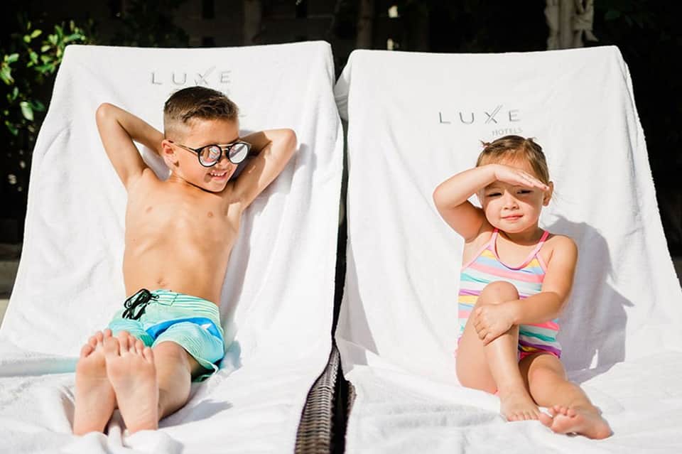 Two children relaxing on white LUXE lounge chairs in bright sunlight by the pool