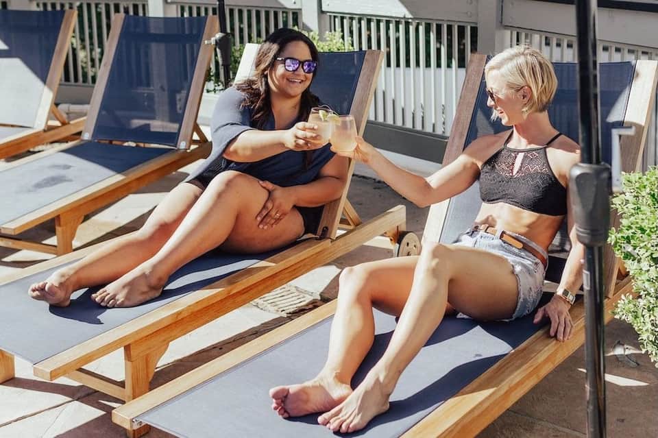 Two women toasting drinks while relaxing on wooden lounge chairs by pool