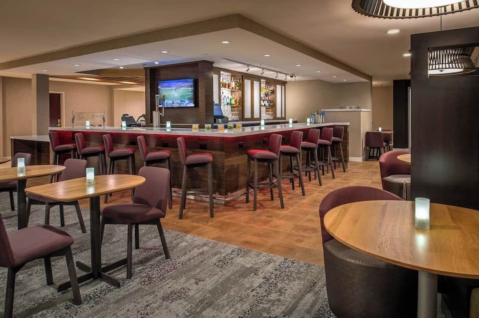 Modern hotel bar and lounge with burgundy seating, wooden tables, and a full-service bar counter with TV screen