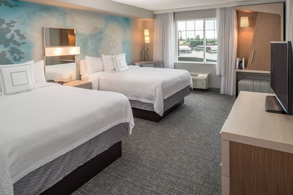 Bright hotel guest room with two queen beds, blue accent wall, river view windows, and modern furnishings