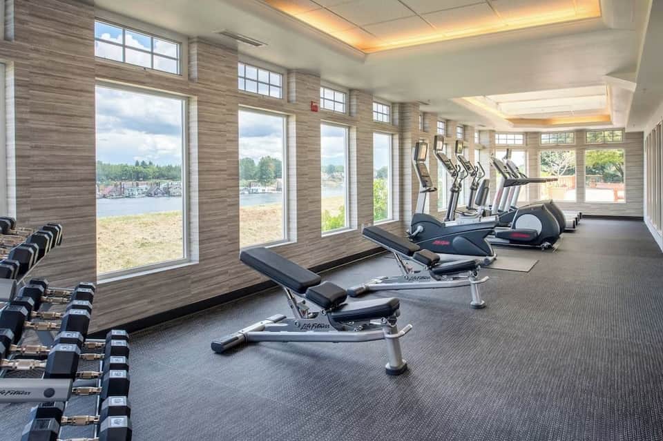 Indoor fitness center with cardio machines, dumbbells, and large windows overlooking waterfront