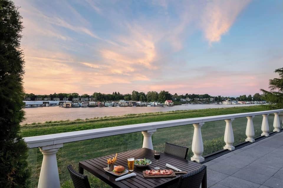 Waterfront balcony dining setup with table, food and drinks, overlooking river at sunset