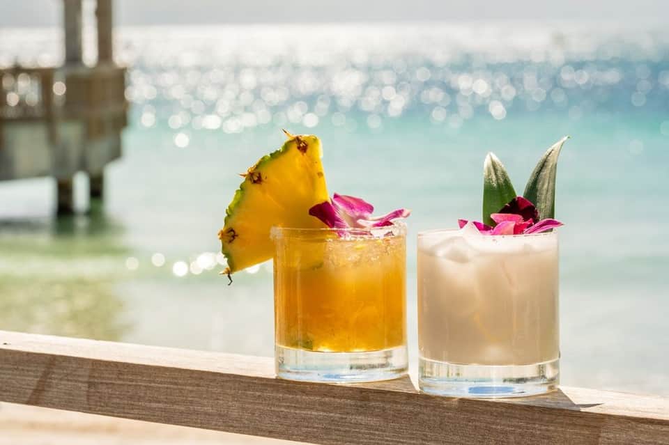 Two tropical cocktails with pineapple and orchid garnish on oceanfront railing overlooking turquoise water