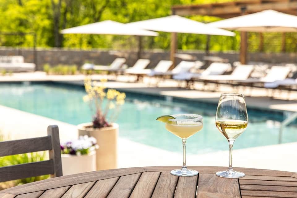 Two white wine glasses on wooden table overlooking resort pool with lounge chairs and tree-lined background