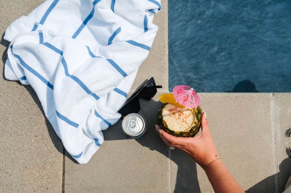 Hand holding pineapple drink with pink umbrella by pool edge with white striped towel nearby