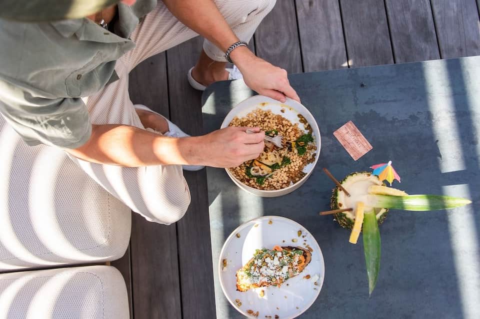 Person dining outdoors holding bowl of quinoa salad with garnished plate and tropical drinks