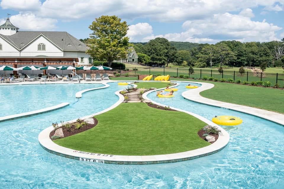 Large resort pool complex with white curved design, grassy islands, yellow floats, clubhouse, and scenic countryside views