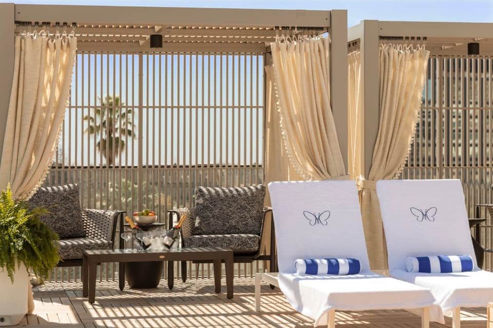 Private pool cabana with beige curtains, comfortable seating, and ocean view through vertical blinds