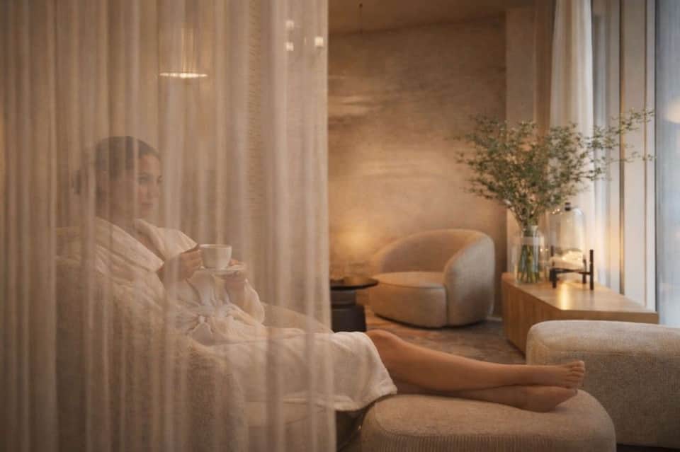 Serene spa lounge with woman holding coffee cup by sheer curtains and curved beige armchair