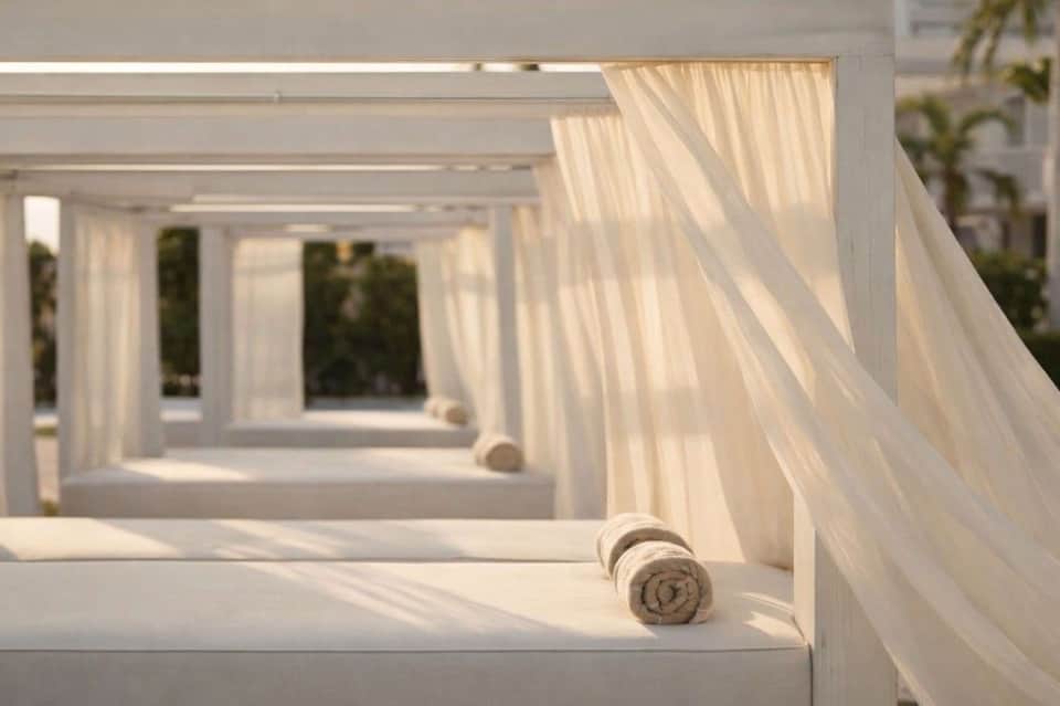 Covered poolside cabana with flowing white curtains, rolled towels, and dappled sunlight