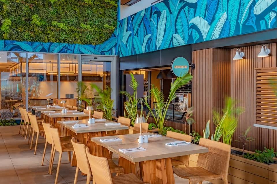 Outdoor restaurant patio with wooden tables, rattan chairs, blue leaf mural, and green living wall
