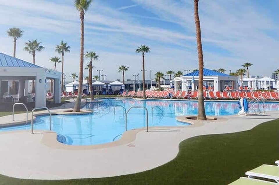 Modern resort pool area with palm trees, red lounge chairs, white buildings, and blue metal roofs