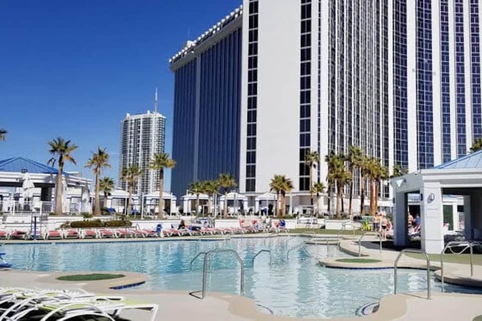 Large resort pool with shallow sections, white cabanas, palm trees, and tall hotel building behind