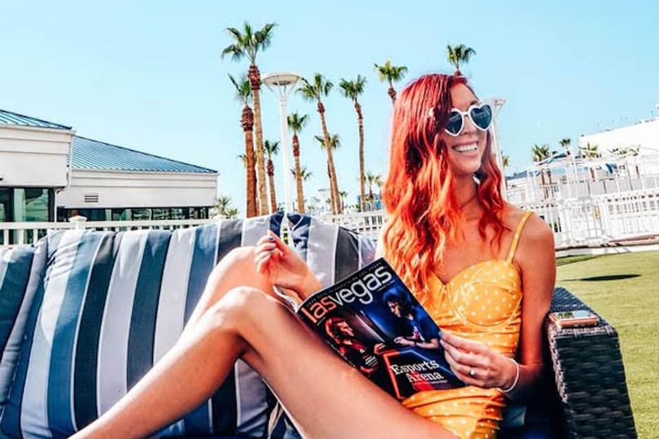 Woman in yellow dress with red hair wearing heart-shaped sunglasses reading magazine by striped lounge chair