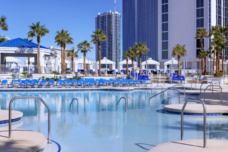 Large beachfront resort pool with blue lounge chairs, palm trees, white umbrellas, and high-rise hotels