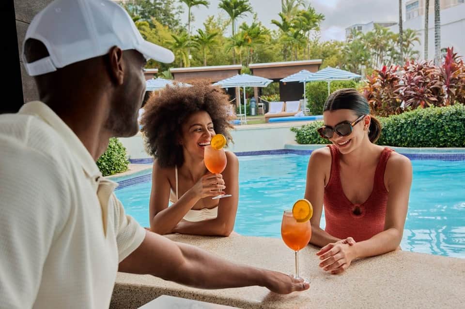 Three guests enjoying cocktails poolside at resort bar with pool in background