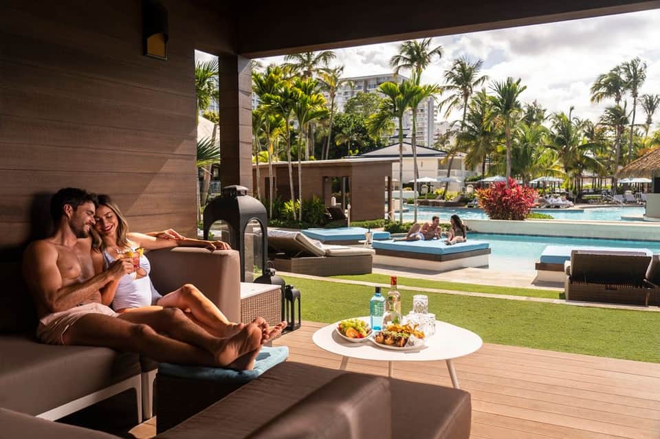 Couple relaxing on covered lounge with view of resort pools, palm trees, and modern architecture