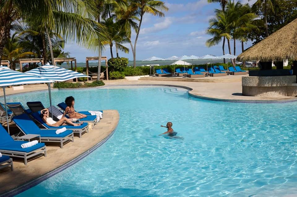 Resort pool with guests relaxing on blue loungers under striped umbrellas, ocean view in background