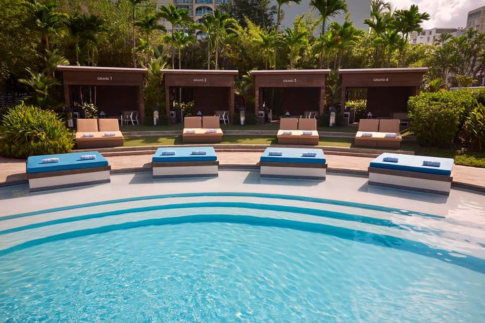 Modern resort pool with curved edges, blue lounge chairs on stone deck, and dark wooden cabanas