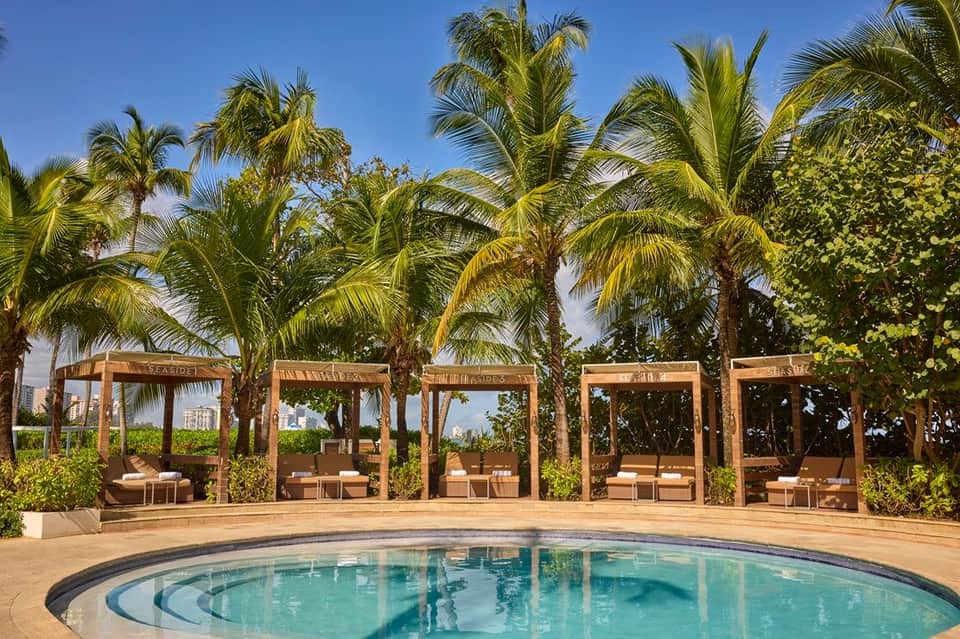Circular resort pool surrounded by wooden cabanas, palm trees, and manicured green lawn areas