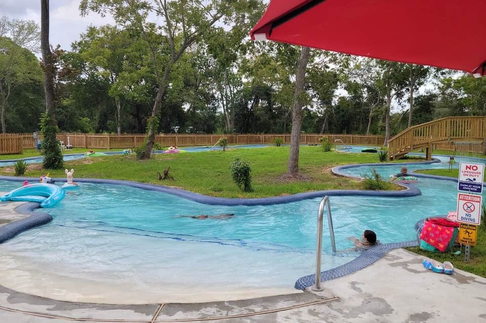 Lazy river pool with turquoise water, sandy beach entry, wooden deck, and families enjoying the water