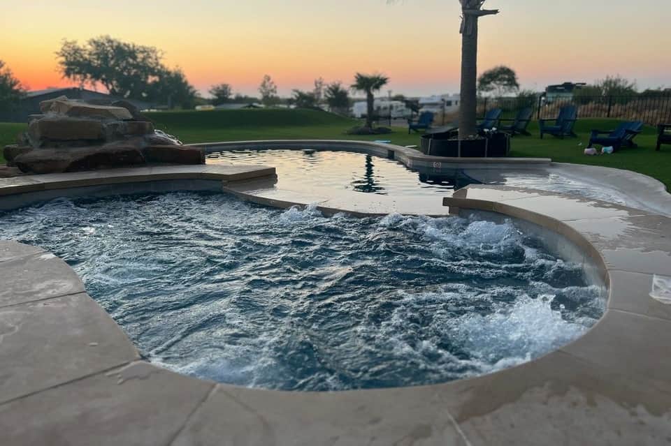 Lit swimming pool with hot tub, waterfall features, natural stone, and lounge chairs at dusk