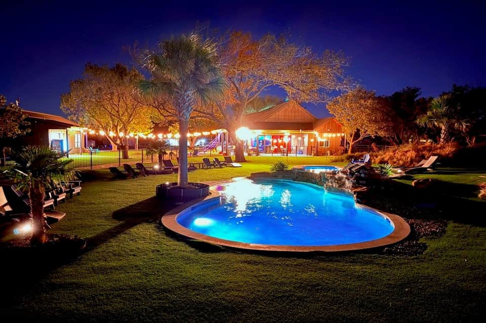 Nighttime resort pool with LED lighting, red buildings with playground, palm trees, and manicured lawn