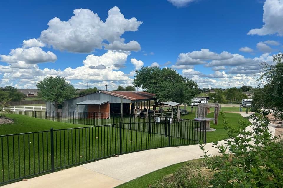 Rural property with metal-roofed barn, black fencing, manicured lawn, and scattered buildings under blue sky with white clouds