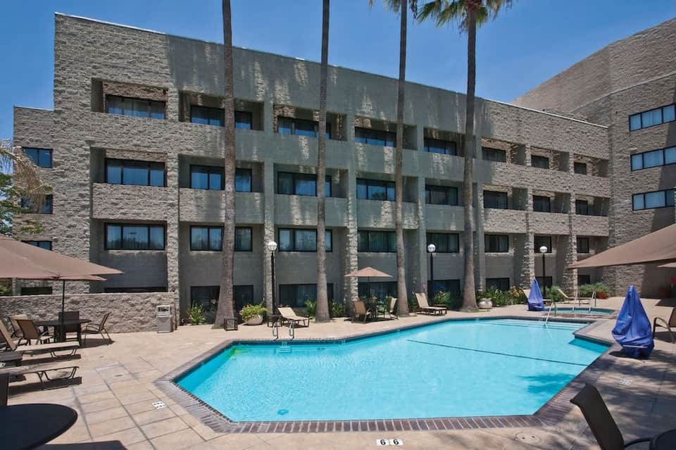 Outdoor swimming pool with lounge chairs, stone building facade, palm trees, and desert landscape