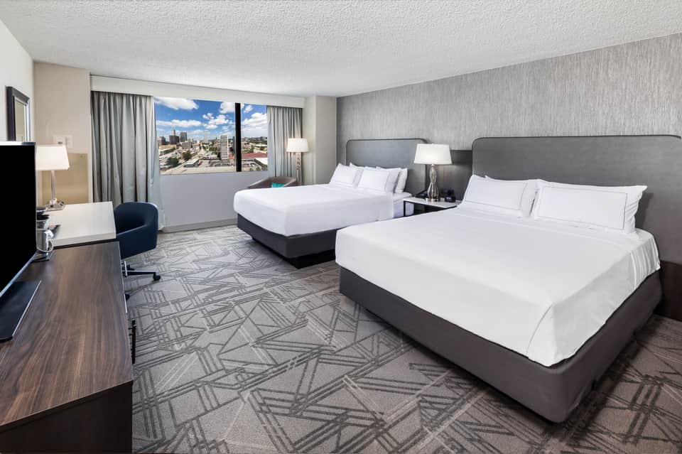 Modern hotel room with two queen beds, city skyline view through windows, work desk, and contemporary décor