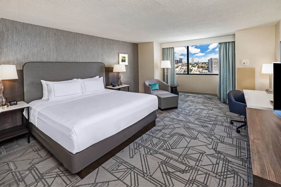 Spacious hotel bedroom with king bed, gray headboard, city view windows, armchair, and geometric patterned carpet