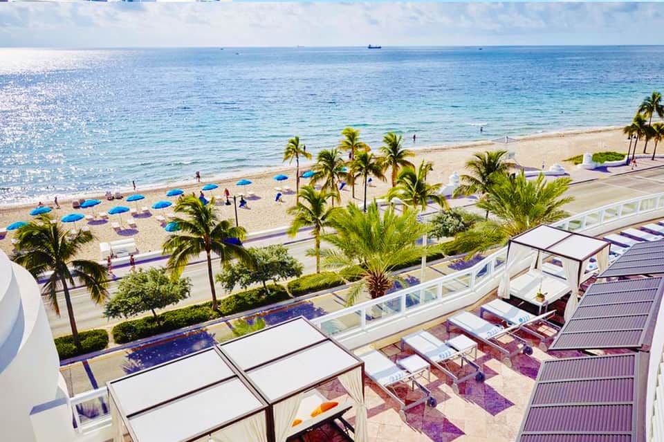 Aerial view of beach with blue umbrellas, palm trees, sandy shore, and ocean with white cabanas
