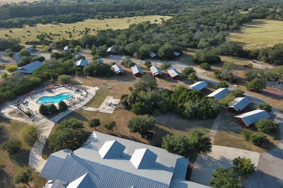 Aerial view of resort with pool, multiple cabin buildings with blue roofs scattered across green pasture