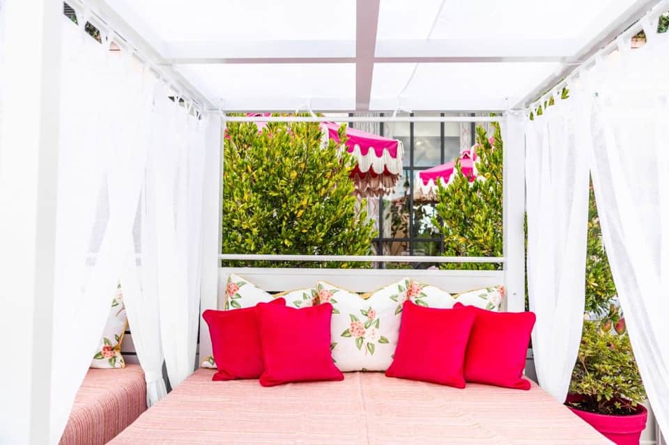 White canopy bed with red pillows and floral bedding on a rooftop terrace, surrounded by boxwood hedges and pink decorative bunting.