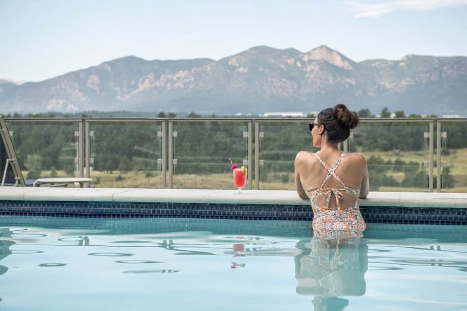 Woman in pool overlooking mountains, holding pink cocktail with glass railing and scenic mountain backdrop