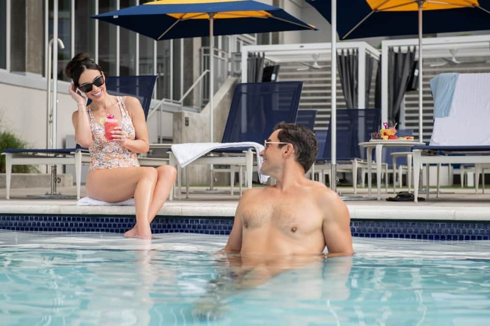 Woman in patterned swimsuit sitting poolside chatting with man in water under large umbrella