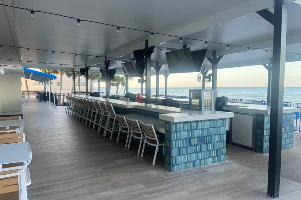 Modern beachfront bar with long counter seating, string lights, and ocean views through open-air covered structure
