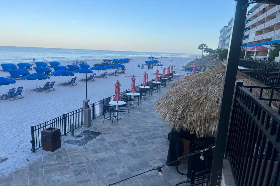 Sandy beach with blue umbrellas and lounge chairs, tiki bar structures, and calm ocean waters at dusk