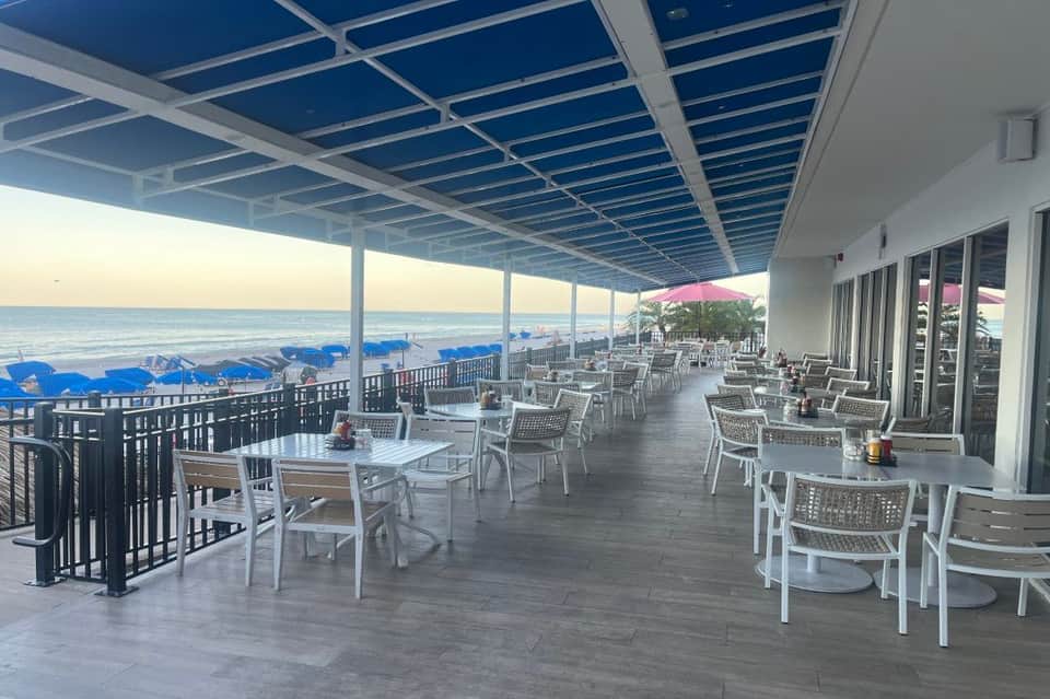 Oceanfront dining patio with white chairs, tables, blue pergola roof, and direct beach access