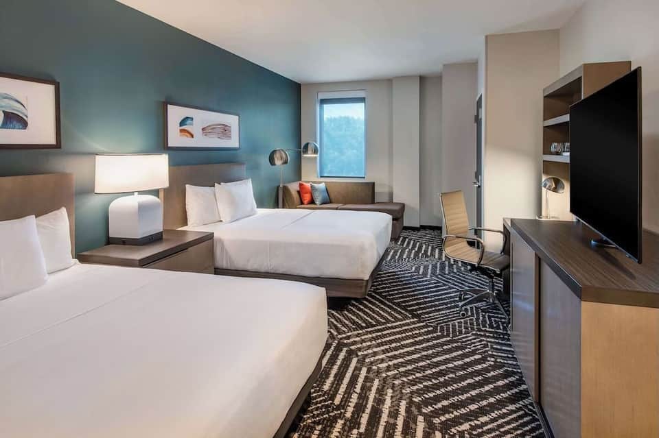 Modern hotel room with two queen beds, teal accent wall, geometric patterned carpet, flat-screen TV, and seating area with window
