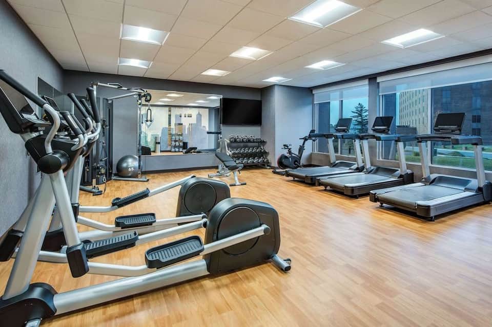 Bright fitness center with exercise bikes, treadmills, weight equipment, and city views through large windows