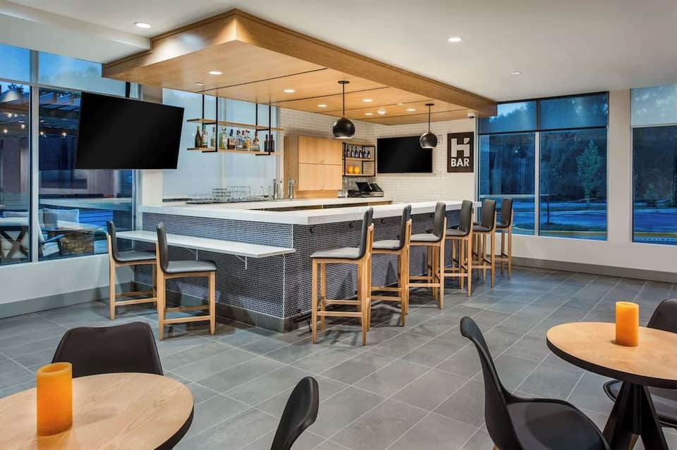 Modern hotel bar lounge with wooden ceiling, white bar counter, seating, flat-screen TVs, and scenic water views